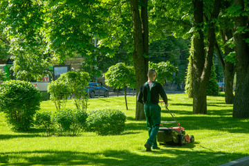 Fototapeta premium Worker with a lawn mower wearing green work suit mows the lawn in the square in the city.