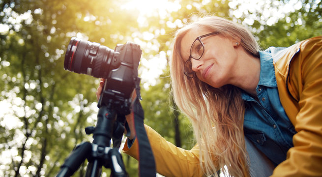 Low angle of blond woman looking at digital camera display taking pictures in green leafy forest