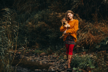 A beautiful young happy woman with long hair holds a small dog with a bow on its head. Autumn. Girl...