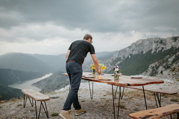 Young man setting up table for picnic outdoors. Mountain top view and canyon in background