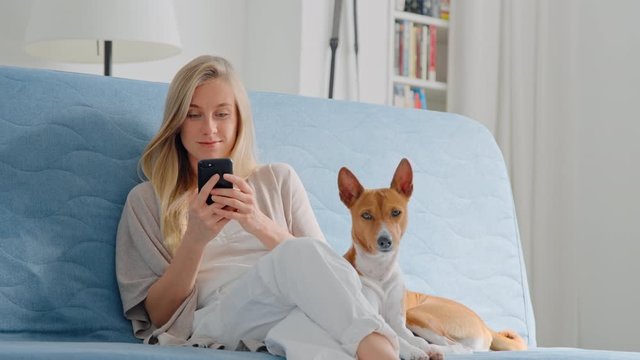 Sweet and adorable young woman sit on sofa, use smartphone and text through social media app. Cute puppy sit with owner woman look into camera and yawn. Work from home and relax in living room