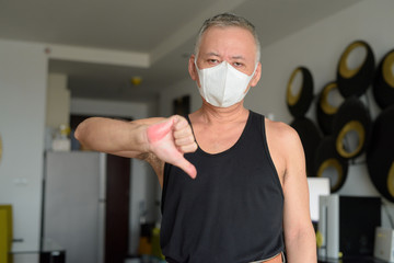 Stressed mature Japanese man with mask giving thumbs down at home under quarantine due to covid-19...