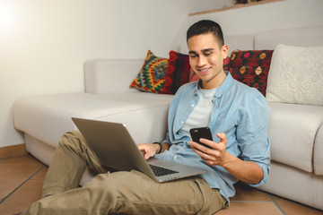 Casual young man at home smiling on computer laptop holding smartphone. Happy for new purchase. Online shopping, internet banking and technology concept.