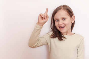 Little smiling girl in a light sweater on a light background raised her hand and shows her thumb up