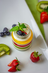A glass jar with the layered multi colors jelly and berries such as blueberries, strawberry, kiwi and mint leaves served on the white plate , green napkin and white pot at the background