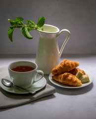 Breakfast table set up with the hot tea and croissant, cheese cubes fresh mint served in white dishes on the gray background.