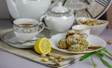Table set up with the home made coconut cookies, white and gray print tea set, black tea, lemon, pistachios nuts, vase with the green branch, wall clocks at the back on the gray background.