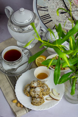 Table set up with the home made coconut cookies, white and gray print tea set, black tea, lemon, pistachios nuts, vase with the green branch, wall clocks at the back on the gray background.