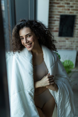 Fototapeta premium Beautiful woman standing in white blanket in light room, morning, relax time at home