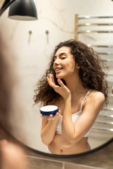 Portrait of happy young woman look in mirror applying cream in bathroom