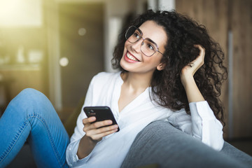 Fototapeta na wymiar Happy pleasant woman relaxing on comfortable couch, holding smartphone in hands. Smiling young lady chatting in social networks, watching funny videos, using mobile applications at home.