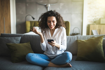Portrait of a suspicious woman holding phone looking at you sitting on a sofa in the living room of...