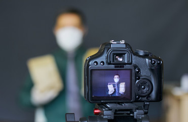 A blogger in a protective mask takes pictures of himself on camera. Advertises a product. Focus is on his camera on a tripod and the image on it. Everything else is blurry.