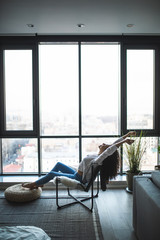 Young woman at home sitting on modern chair in front of window relaxing in her living room