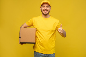 Portrait of excited delivery man in yellow uniform holding paper box isolated over yellow...