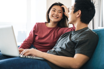 Fototapeta premium Photo of asian couple laughing and using laptop while sitting on sofa