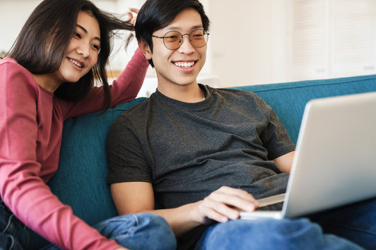 Photo of asian couple laughing and using laptop while sitting on sofa