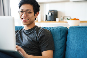 Photo of cheerful asian man using laptop while sitting on sofa