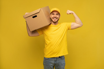 Portrait of excited delivery man in yellow uniform holding paper box isolated over yellow...