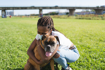 African woman with braids and protective mask embracing a boxer dog outside