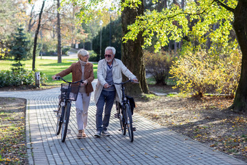 Obraz premium Mature fit couple pushing bicycles in public park talk and smile