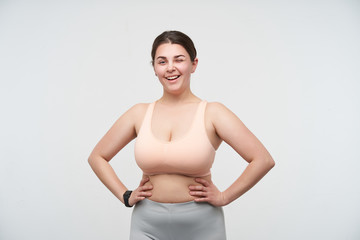 Fototapeta na wymiar Cheerful young dark haired plump lady in sports bra and leggins keeping hands on waist and winking happily at camera while standing over white background