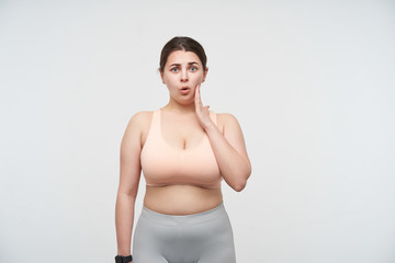 Fototapeta na wymiar Bewildered young dark haired chubby woman with ponytail hairstyle holding raised palm on her cheek while looking confusedly at camera, isolated over white background