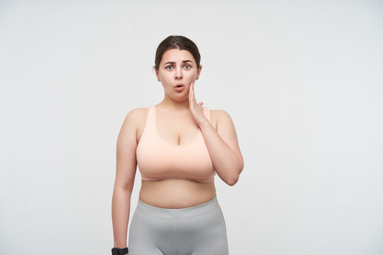 Bewildered young dark haired chubby woman with ponytail hairstyle holding raised palm on her cheek while looking confusedly at camera, isolated over white background