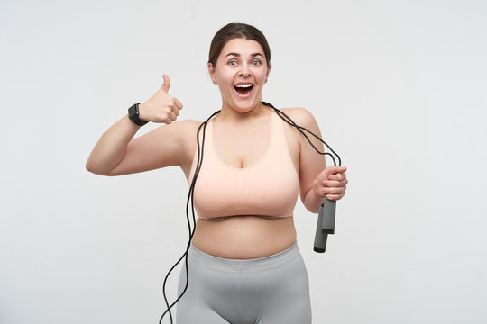 Agitated young dark haired plump woman dressed in sporty clothes wearing jumping-rope and showing happily thumb to camera while posing over white background. Concept of weight loss