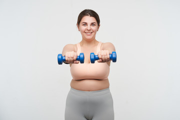 Studio shot of young pleased oversized female making static exercises with weight and smiling...