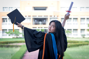 Portrait of happy and excited of young Asian female university graduate wears graduation gown and hat celebrates with degree in university campus in the commencement day. Education concept.