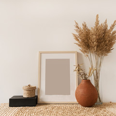 Blank photo frame, fluffy reeds bouquet in vase, rye in clay pot at white wall. Copy space mockup...