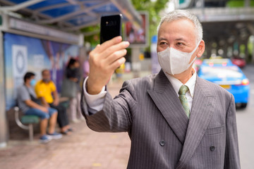 Mature Japanese businessman with mask taking selfie at the bus stop