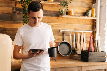 Portrait of young happy smiling man in casual clothes holding digital tablet and coffee cup or tea in cozy kitchen room with wooden walls.