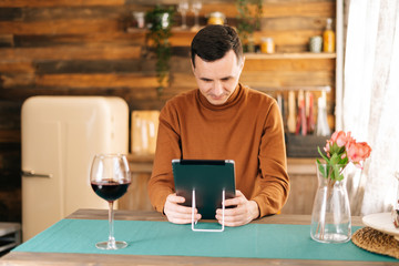 Young happy man is using digital tablet, high glass with red wine on the table in cozy kitchen room with wooden walls.