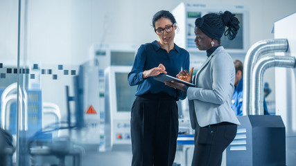 Obraz premium Industrial Engineer Talks with Project Supervisor, They Use Digital Tablet Computer and Discuss Modern Factory Planning. Manufacturing Facility with CNC Machinery and Robot Arm Assembly Line