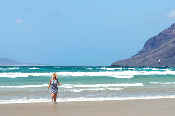 Blonde caucasian white woman in blue short dress walking and enjoying sunny day In Famara beach in the northwest of the island of Lanzarote,one of the most popular places to surf in the Canary Islands