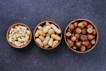 Three wooden bowls with hazelnuts, cashews and peanuts on grey table, top view