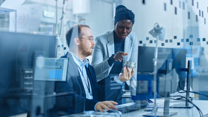 Modern Factory: Female Project Supervisor Talks to a Male Industrial Engineer who Works on Computer. They Inspect Machinery Mechanism and Design Improved Version. Working High-Tech Industrial Facility