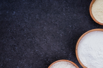 Dark marble background with rice flour, corn flour and whole grain flour in wooden bowls. Baking ingredients with space for text or image. Flat lay top view, studio shot