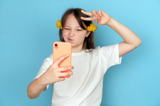 Portrait of a girl with dandelions in her hair and with a phone in her hands on a blue background. Studio photo. Selfie concept and blogger. Emotion of contempt and the sign of Victoria.