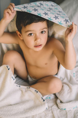 overhead view of cute boy, wrapped in hooded towel, sitting on bed and looking at camera