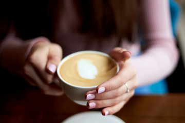Fototapeta na wymiar Close-up picture of female hands with pink manicure, holding white coffee mug with cappuccino foamy hot drink. Sunday leisure time. Food and drink establishment.