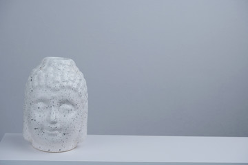 white Buddha head statuette on a white table against gray concrete wall copy space
