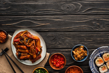 top view of korean topokki near kimbap, chopsticks, side dishes and cotton napkin on wooden surface