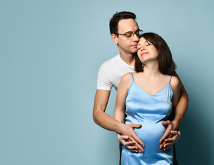 Naklejka premium Pregnant female in blue nightgown. Husband hugging her, they looking at each other, put hands on her belly posing on blue background
