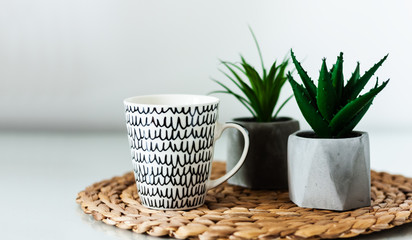 Cozy home interior decor: white and black cup and ornamental plants in pots on a wicker stand on a white table in the room. The quarantine concept of stay home.