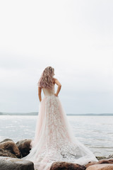 Fototapeta na wymiar A girl in a wedding dress stands on the rocks, her back facing forward, on the beach.in a soft pink long dress
