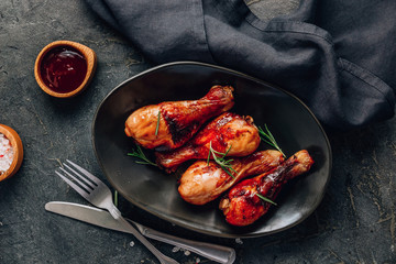 Chicken legs on black plate with red sauce and rosemary on black rustic background.
