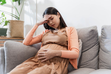 Obraz premium Young pregnant asian woman has suffering from headache sitting on sofa. Pregnancy symptoms